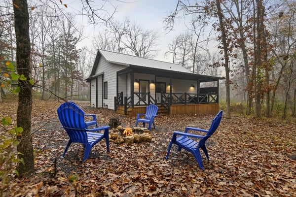 Modern cabin with white siding and metal roof surrounded by bare trees, blue Adirondack chairs around fire pit in leaf-covered yard.