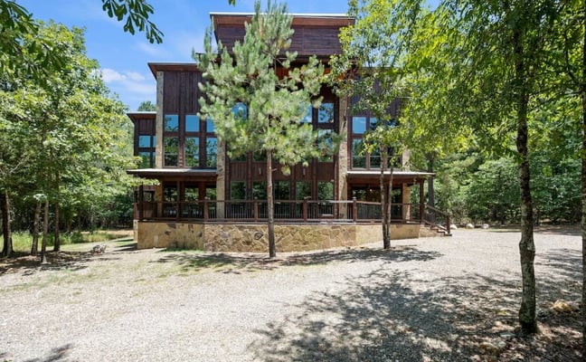 Modern multi-story house with brick and wood exterior, large windows, and wraparound deck surrounded by tall trees